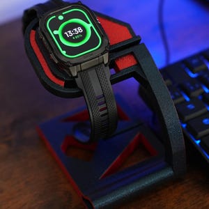 Puede incluir: Un reloj inteligente negro con una correa negra se muestra en un soporte negro y rojo. La pantalla del reloj muestra la hora 13:38 y una carga de batería del 95%. El soporte está sobre una superficie de madera.