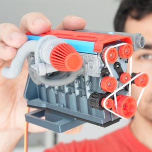 Puede incluir: Un modelo detallado de un motor, con una parte superior roja y azul, componentes grises y detalles en naranja. El modelo incluye un turbocompresor, poleas y una correa, mostrando detalles mecánicos intrincados. El motor se sostiene en una mano.