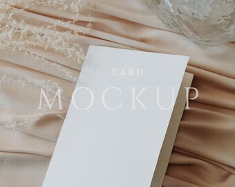 May include: A white card mockup with the word "MOCKUP" and the word "CARD" printed on the front. The card is open, revealing the inside. The card is on a peach-colored fabric with a glass vase and dried flowers.