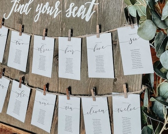 May include: A rustic wooden sign with the text "Find your seat" written in white calligraphy.  Cards with table numbers and guest names are attached to the sign with clothespins and twine.