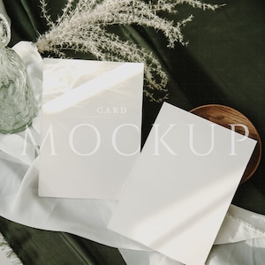 May include: A mockup of two white cards with the word "CARD" and "MOCKUP" printed on them. The cards are on a white cloth, with a dark green background. A wooden plate and dried flowers are also in the image.