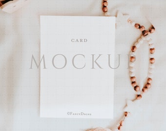 May include: A white card mockup with the text "CARD MOCKUP" and the copyright symbol followed by "FANCYDSGNS". The card is on a white textured surface with a brown and white beaded garland.