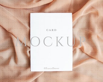 May include: A white card mockup on a peach-coloured fabric background. The card has the text "CARD MOCKUP" and the copyright symbol with "FANCYDESIGNS".