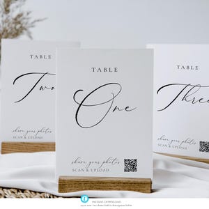 Op de afbeelding: Drie witte tafelnummerkaarten met elegant zwart schrift. Elke kaart wordt op een houten voet gepresenteerd. De kaarten luiden "Table One", "Table Two" en "Table Three". De kaarten bevatten ook de tekst "share your photos SCAN & UPLOAD" en een QR-code.