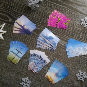 May include: A collection of eight different cards with various nature scenes. The cards feature images of a sunset, a beach with a cross, a flock of birds, a cloudy sky, and a field of pink flowers.