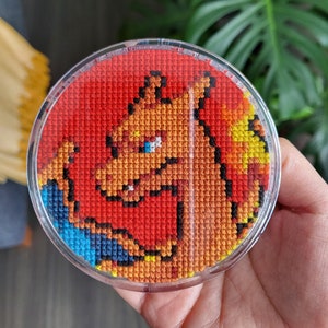 May include: A round, clear plastic frame with a red, orange, and yellow cross-stitch design of a cartoon character. The character has a red body, orange mane, and yellow eyes.