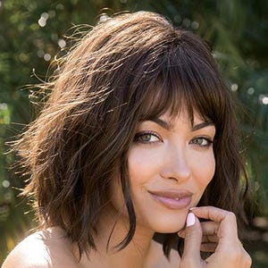 May include: A woman with brown hair and bangs, wearing a yellow off-the-shoulder top. The woman is smiling and looking towards the camera. The background is blurred with green foliage.