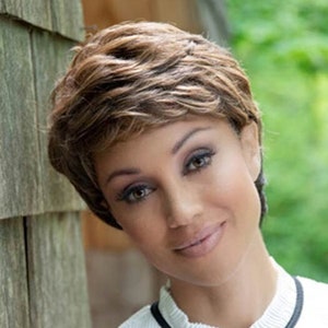 May include: A woman with short, wavy, brown hair is wearing a white shirt with a black collar and a black bow tie. She is standing next to a wooden wall.