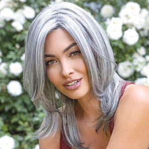 May include: A woman with a medium-length, layered gray wig. The wig has a natural-looking hairline and is styled with soft waves. She is wearing a burgundy tank top. White flowers are in the background.