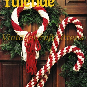 Puede incluir: Una corona de Navidad roja y blanca de ganchillo con un bastón de caramelo rojo y blanco de ganchillo colgado de una puerta de madera. La corona y el bastón de caramelo están decorados con borlas blancas. El texto "Yuletide Greetings" está escrito en letras amarillas en la puerta.