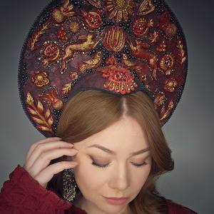 Könnte beinhalten: Ein dekorativer, verzierter Kopfschmuck mit dunkelroter Basis, verziert mit goldener und roter Stickerei. Das Design umfasst Blumen- und Tiermotive. Der Kopfschmuck wird von einer Person getragen, die einen roten Pullover trägt.