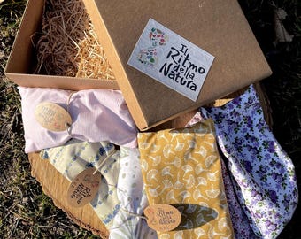 May include: A brown cardboard box with a label that reads "Il Ritmo della Natura" contains several fabric-covered items. The items are in various colors and patterns, including pink stripes, yellow with white leaf designs, and purple floral.