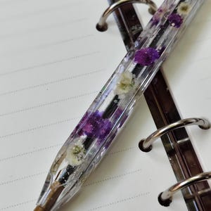 Könnte beinhalten: Ein klarer Harzstift mit kleinen lila und weißen Blumen. Der Stift liegt auf einem linierten Notizbuch mit silbernen Binderingen. Die Spitze des Stifts ist bronzefarben.