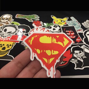 May include: A red and yellow Superman logo sticker with dripping blood. The sticker is on a black background with other stickers around it.