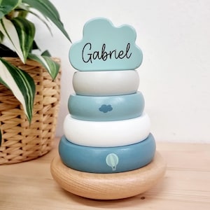 Può includere: Un giocattolo di impilamento in legno blu e bianco con una cima a forma di nuvola che dice "Gabriel". Il giocattolo ha disegni di mongolfiera e nuvola sugli anelli.