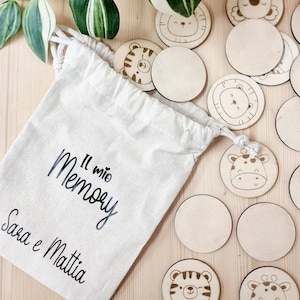 Può includere: Una borsa di tela bianca con la scritta "Il mio Memory Sara e Mattia" stampata su di essa. La borsa contiene un set di pezzi di gioco di memoria in legno con illustrazioni di animali, tra cui tigri, giraffe e un koala.