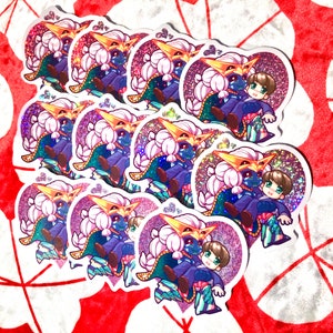 May include: A stack of heart-shaped stickers featuring a cartoon character with three eyes, pink hair, and a blue outfit. The character is holding a smaller character with brown hair. The stickers have a glittery finish.