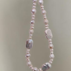 May include: A delicate necklace featuring a mix of white and off-white pearls. The necklace includes various sizes and shapes of pearls, including some larger, irregularly shaped baroque pearls. The clasp appears to be gold.