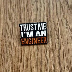 Puede incluir: Pin de esmalte negro y naranja con el texto "Trust me I'm an Engineer".