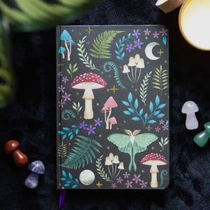 May include: A black journal with a colorful illustration of mushrooms, leaves, and a moth on the cover. The illustration is on a dark background with gold accents.