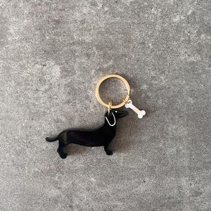 Puede incluir: Llavero de dachshund negro con un anillo dorado y un abalorio de hueso blanco.
