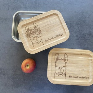 Könnte beinhalten: Zwei Holz-Lunchboxen mit einem Lama-Design und dem Text "Hier kommt ein Alpaka" eingraviert. Eine Lunchbox ist geschlossen und die andere ist offen und zeigt einen Edelstahlbehälter im Inneren.