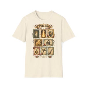 May include: Cream-colored t-shirt featuring a stamp-like design with the word "Paladin" at the top. The design includes images of a sword, a flame, a shield, a horse, a helmet, a compass, a hammer, and a spear.