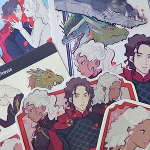 May include: A collection of stickers and art prints featuring fantasy characters with white hair and dark-haired figures in red and black attire, alongside dragon illustrations. Some stickers are labeled with the text Prince and Team Black.