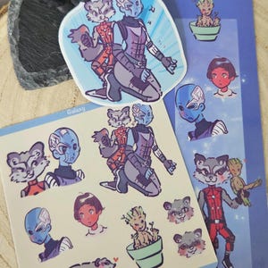 Può includere: Una collezione di adesivi e un segnalibro con illustrazioni a cartoni animati di personaggi, tra cui un procione, un umanoide dalla pelle blu e una piccola creatura vegetale. I disegni includono ritratti, un procione in tuta rossa e una creatura in un vaso, con le scritte Galaxy e raccoon stampate.