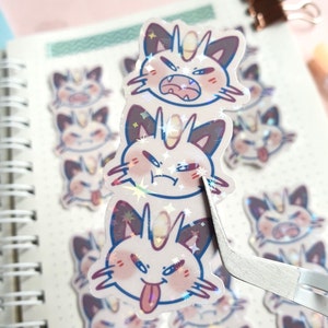 May include: Three holographic stickers featuring a cartoon cat with different facial expressions. The cat has pink cheeks, a white horn, and a pink tongue sticking out in one of the stickers.