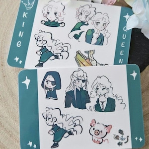 May include: Two teal-colored sticker sheets with illustrations of cartoon characters. One sheet reads "KING" and the other reads "QUEEN". The stickers are on a wooden surface with flowers.