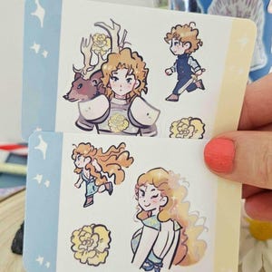 以下が含まれることがあります： カールした髪、鎧、花のアクセントが特徴のイラストキャラクターが描かれた2枚のステッカーシート。1枚のステッカーには鹿とキャラクターが、もう1枚には剣とキャラクターが描かれています。ステッカーは水色と黄色のボーダーです。