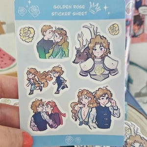 以下が含まれることがあります： 「GOLDEN ROSE STICKER SHEET」の文字と、カップルや鎧姿のキャラクターのイラストが描かれたステッカーシート。ステッカーは水色の縁取りと花の装飾が特徴です。シートは人が持っています。