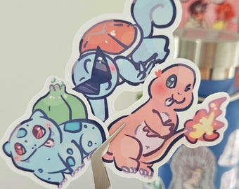 May include: A close-up of three cartoon stickers featuring popular characters. The stickers are held by tweezers. The characters are a blue turtle, a pink lizard with a flame, and a green plant-like creature.