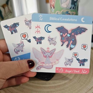 May include: Two sticker sheets featuring various cartoon creatures with wings and heart symbols. One sheet has the text "Biblical Eeveelutions" and "Angel / Devil". The stickers are in shades of blue, pink, and purple.