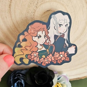 May include: A sticker featuring two cartoon women with long hair, one red and one white, surrounded by orange flowers. The women are wearing dark green and blue dresses. The sticker is on a wooden surface with a hand holding it, and flowers are in the background.