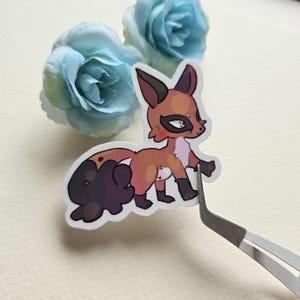 May include: A sticker featuring a cartoon fox with orange fur, black markings, and a white chest. The fox is standing on its hind legs with its tail curled around its body.