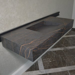 Può includere: Un piano di lavoro in marmo grigio scuro con un'incisione rettangolare per un lavabo. Il piano di lavoro presenta un motivo di venature dorate ed è fissato a una parete con una staffa in metallo nero.