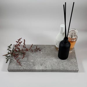 May include: A rectangular gray stone tray with a textured surface. A black diffuser bottle with reeds, two perfume bottles, and a sprig of dried flowers are arranged on the tray. The bottles are white, black, and clear.
