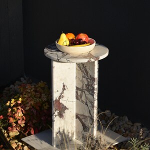 May include: A marble side table with a round top and a rectangular base. A bowl of fruit, including a yellow pear, orange, and red apple, sits on the table. The table has a white and burgundy pattern.
