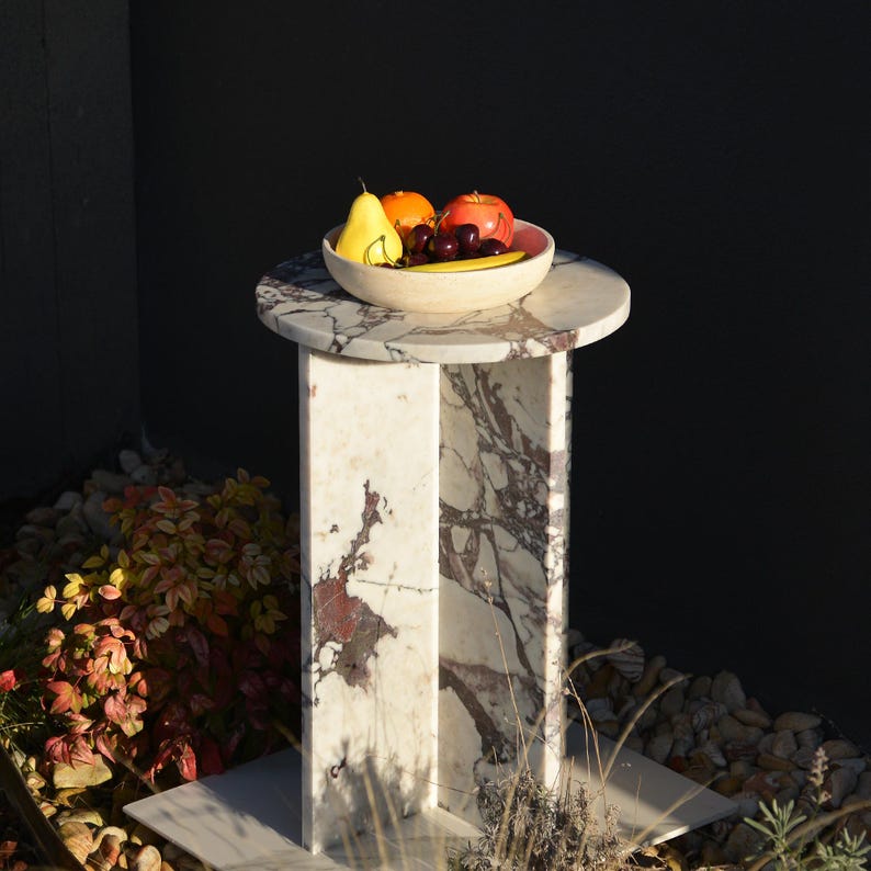 May include: A marble side table with a round top and a rectangular base. A bowl of fruit, including a yellow pear, orange, and red apple, sits on the table. The table has a white and burgundy pattern.