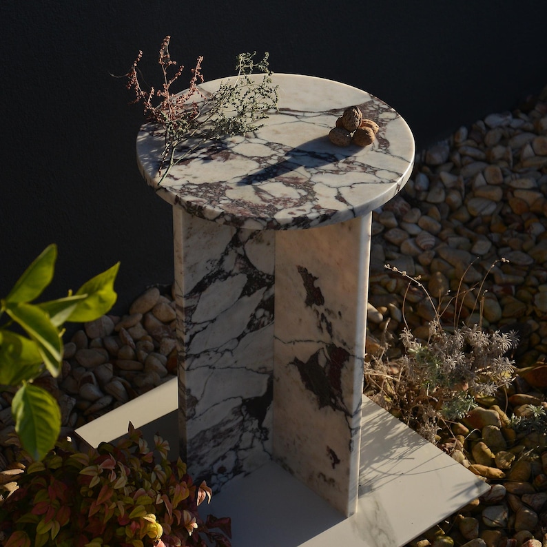 May include: A round marble side table with a white and purple veined pattern. The table has a single rectangular support and a square base. The table is decorated with small plants and a few walnuts.