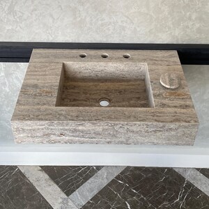 May include: A rectangular, stone bathroom sink with a light brown and gray color scheme. The sink has a rectangular basin, three faucet holes, and a circular soap dish. The sink is mounted on a wall.