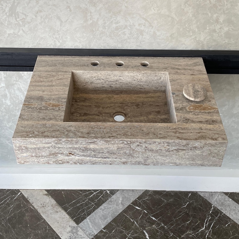 May include: A rectangular, stone bathroom sink with a light brown and gray color scheme. The sink has a rectangular basin, three faucet holes, and a circular soap dish. The sink is mounted on a wall.