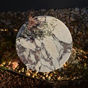 May include: A round marble table with a white and burgundy veined pattern. The table is topped with dried flowers and sits on a bed of small stones. The table's diameter is approximately 24 inches.