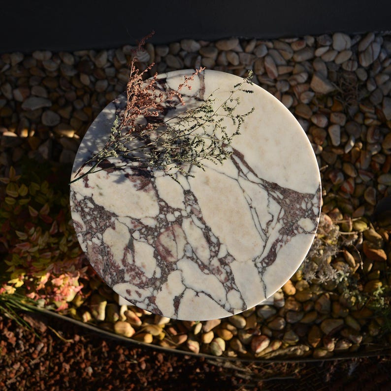 May include: A round marble table with a white and burgundy veined pattern. The table is topped with dried flowers and sits on a bed of small stones. The table's diameter is approximately 24 inches.