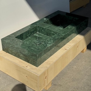 Peut inclure: Un évier rectangulaire en marbre vert avec un bassin et un drain intégrés. L'évier est posé sur une caisse en bois de couleur claire. Le marbre présente un veinage naturel et le design de l'évier est moderne et minimaliste.
