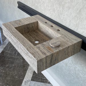 May include: A rectangular, wall-mounted bathroom sink made of gray and brown stone. The sink has a rectangular basin, a circular drain, and three small holes for fixtures. A matching stone disc is on the right side.