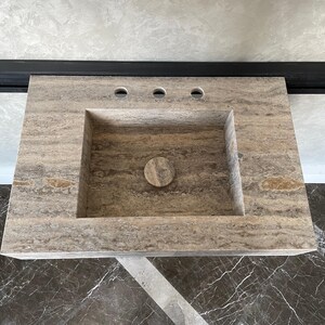 May include: A rectangular, stone bathroom sink with a built-in basin and three faucet holes. The sink is a neutral beige color with natural veining and a circular drain. The sink is designed for countertop installation.