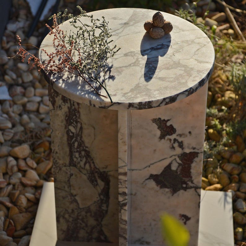 May include: A marble pedestal table with a round top and a cylindrical base. The table features a natural stone pattern with shades of white, gray, and burgundy. A small arrangement of nuts sits on top, with a sprig of dried plants.